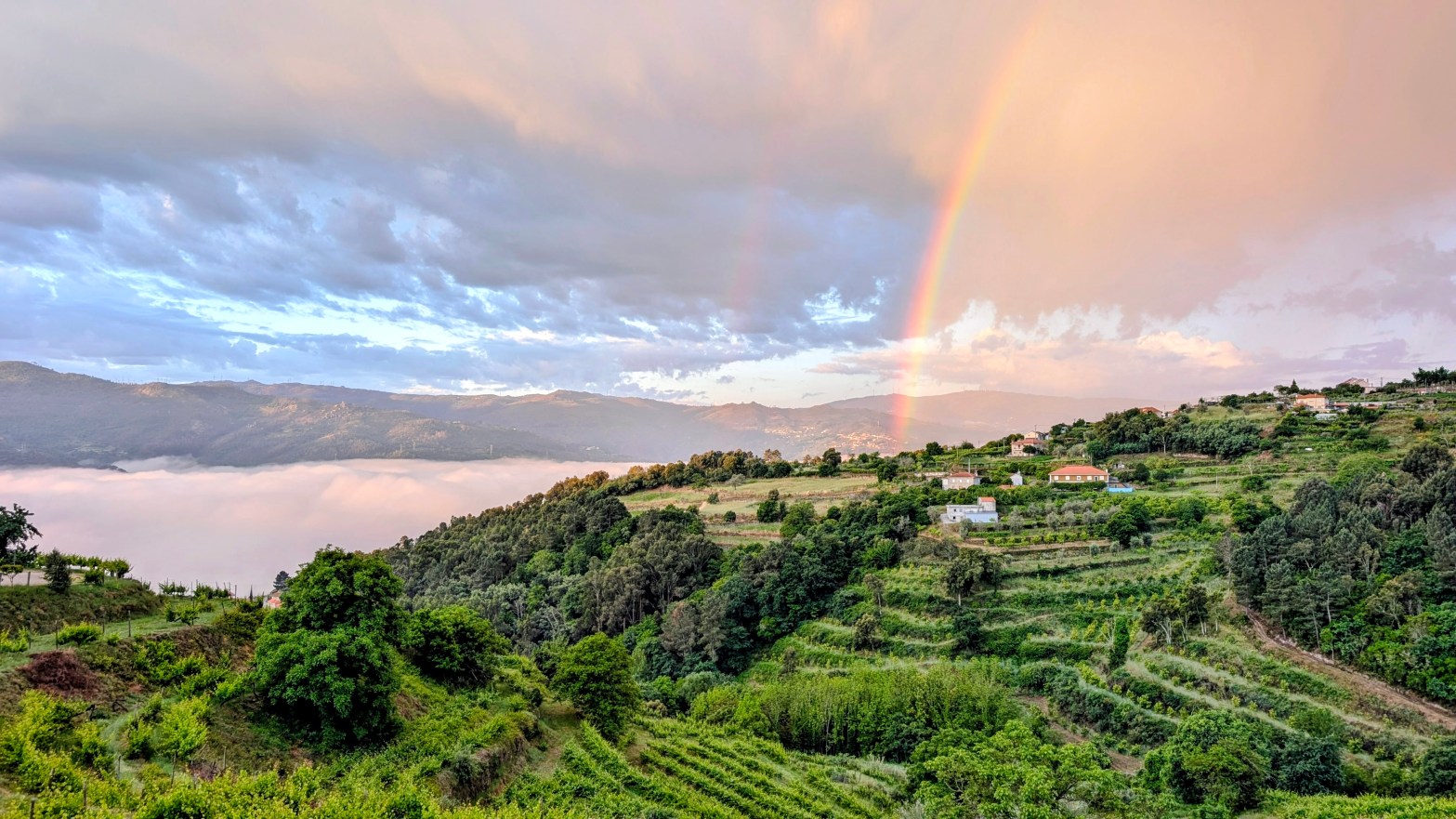 Nascer do dia, as nuvens preenchem o vale do Douro e à direita há uma arco-íris que termina numa pequena aldeia.