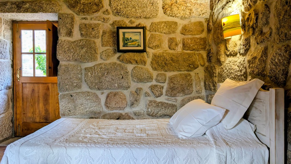 Quarto em casa de pedra com cama feita e a porta de madeira
